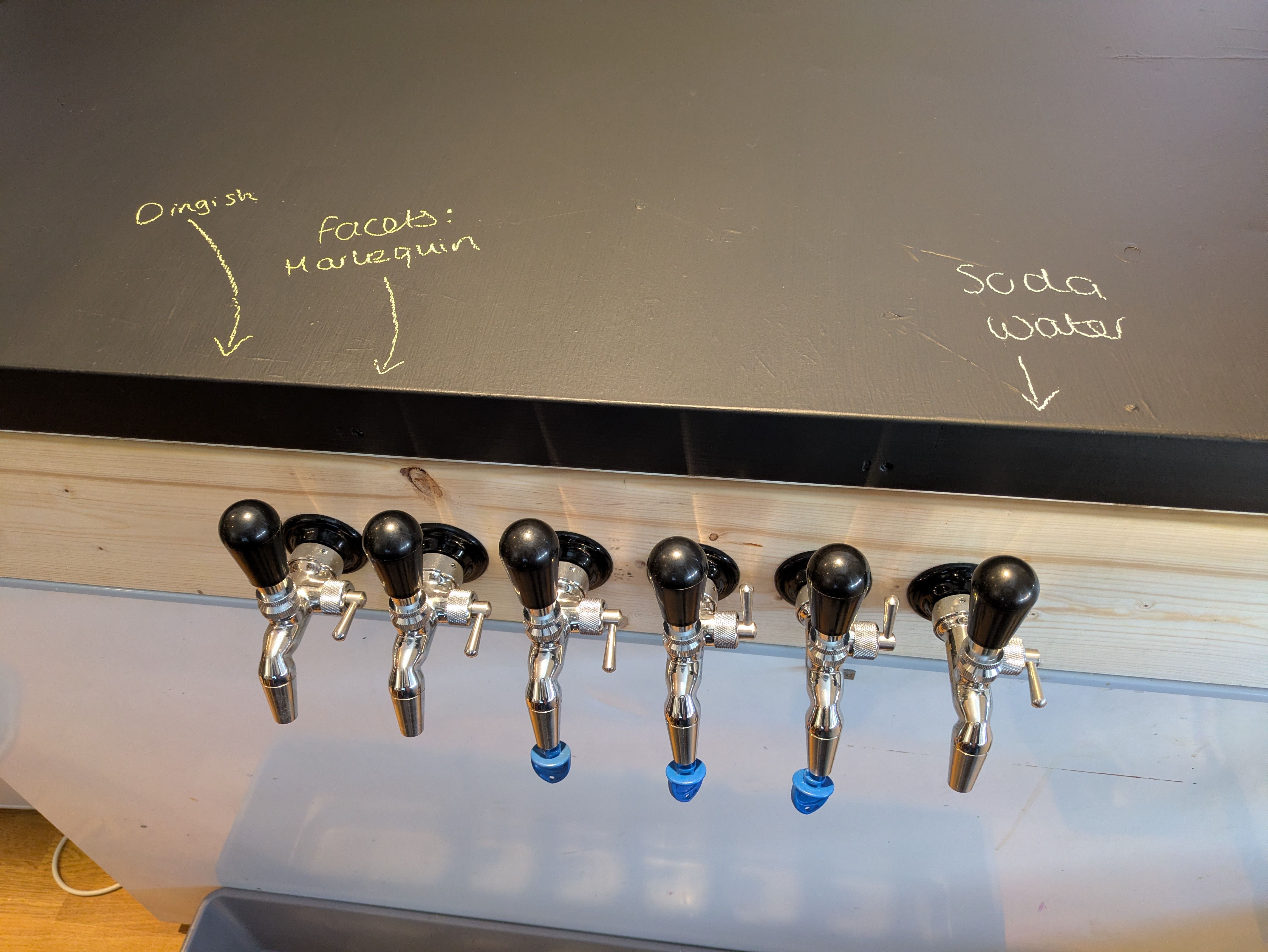 Lid of the keezer, painted with blackboard paint. Written in chalk are the names of the first three beers, with arrows pointing at the relevant taps.